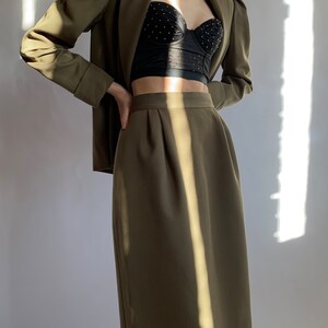 May include: A green blazer with puffed sleeves and a matching green skirt. The blazer is open and a black lace bra is visible underneath.