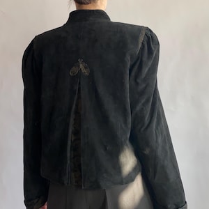 Op de afbeelding: Een zwart su&egrave;de jack met een hoge kraag en pofmouwen. Het jack heeft een decoratief detail op de rug.