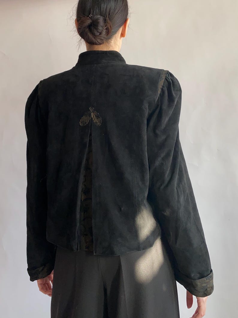 Op de afbeelding: Een zwart su&egrave;de jack met een hoge kraag en pofmouwen. Het jack heeft een decoratief detail op de rug.