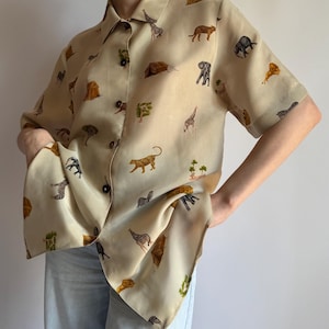 Op de afbeelding: Een overhemd met korte mouwen en knoopsluiting in een lichtbeige kleur, voorzien van een all-over print van diverse Afrikaanse wilde dieren zoals giraffen, olifanten, zebra's en luipaarden, samen met kleine bomen en tenten, afgewerkt met donkere knopen.