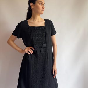 May include: Black vintage dress with a square neckline and short sleeves. The dress features a textured panel down the front with a bow detail at the waist. The dress is knee-length and has a simple, classic design.