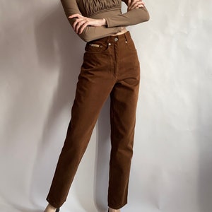 May include: A brown long-sleeved top with a gathered neckline and brown high-waisted jeans. The model is wearing black flats.