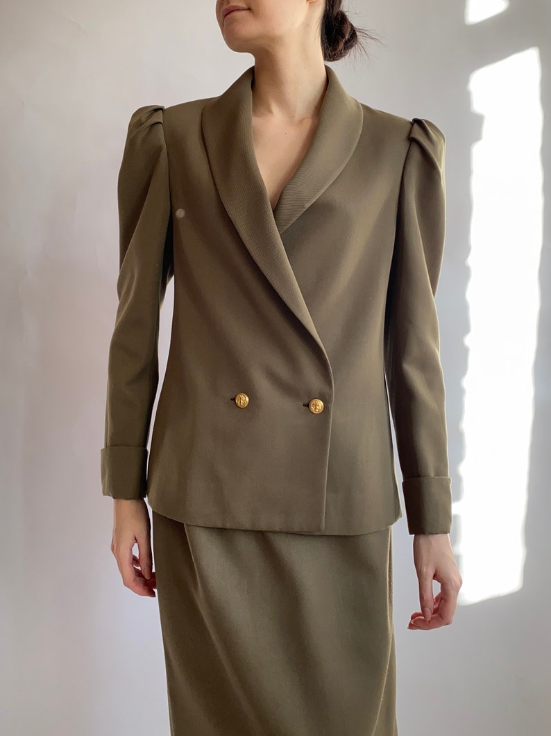 May include: A double-breasted olive green blazer with a wide collar and puffed shoulders. The blazer has gold buttons and a matching skirt.