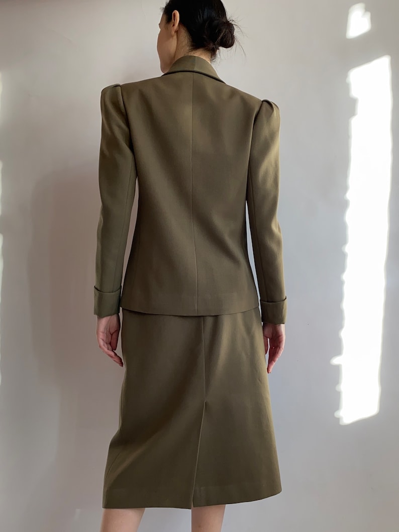 May include: A brown skirt suit with a blazer featuring puffed shoulders and a skirt that hits just above the knee.