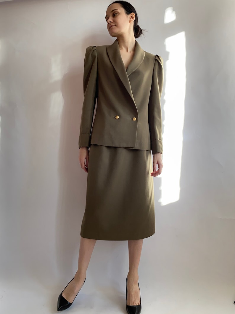 May include: A two-piece suit in a muted olive green colour. The jacket features a double-breasted closure with gold buttons and puffed shoulders. The skirt is knee-length and has a straight silhouette.