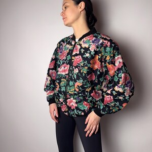 Op de afbeelding: Zwarte bomberjack met een levendig bloemenpatroon met roze, oranje en paarse bloemen. De jas heeft een zwarte kraag, manchetten en ritssluiting. De mouwen zijn licht gezwollen.