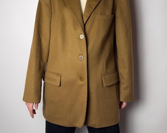 May include: A tan blazer with a classic design. The blazer has a notched lapel, three buttons, and two front pockets. The sleeves are long, and the garment appears to be made of a wool blend. The blazer is worn over a white top and black pants.