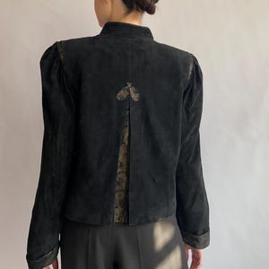 Op de afbeelding: Een zwart su&egrave;de jack met een gouden bloemenpatroon op de rug. Het jack heeft een staande kraag en lange mouwen.