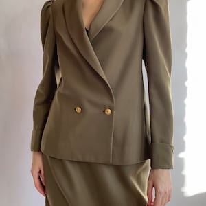May include: A double-breasted blazer with a wide collar and puffed sleeves. The blazer is a dark olive green colour and has gold buttons. The blazer is paired with a matching skirt.