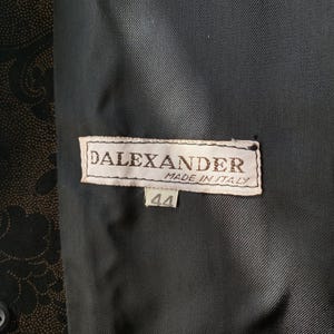 Op de afbeelding: Een zwart-wit label met de tekst "DALEXANDER MADE IN ITALY" en het nummer "44" op een zwarte stoffen achtergrond.