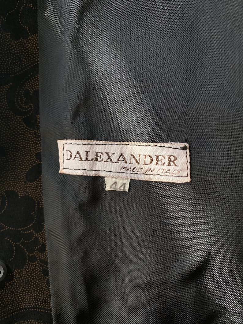 Op de afbeelding: Een zwart-wit label met de tekst "DALEXANDER MADE IN ITALY" en het nummer "44" op een zwarte stoffen achtergrond.