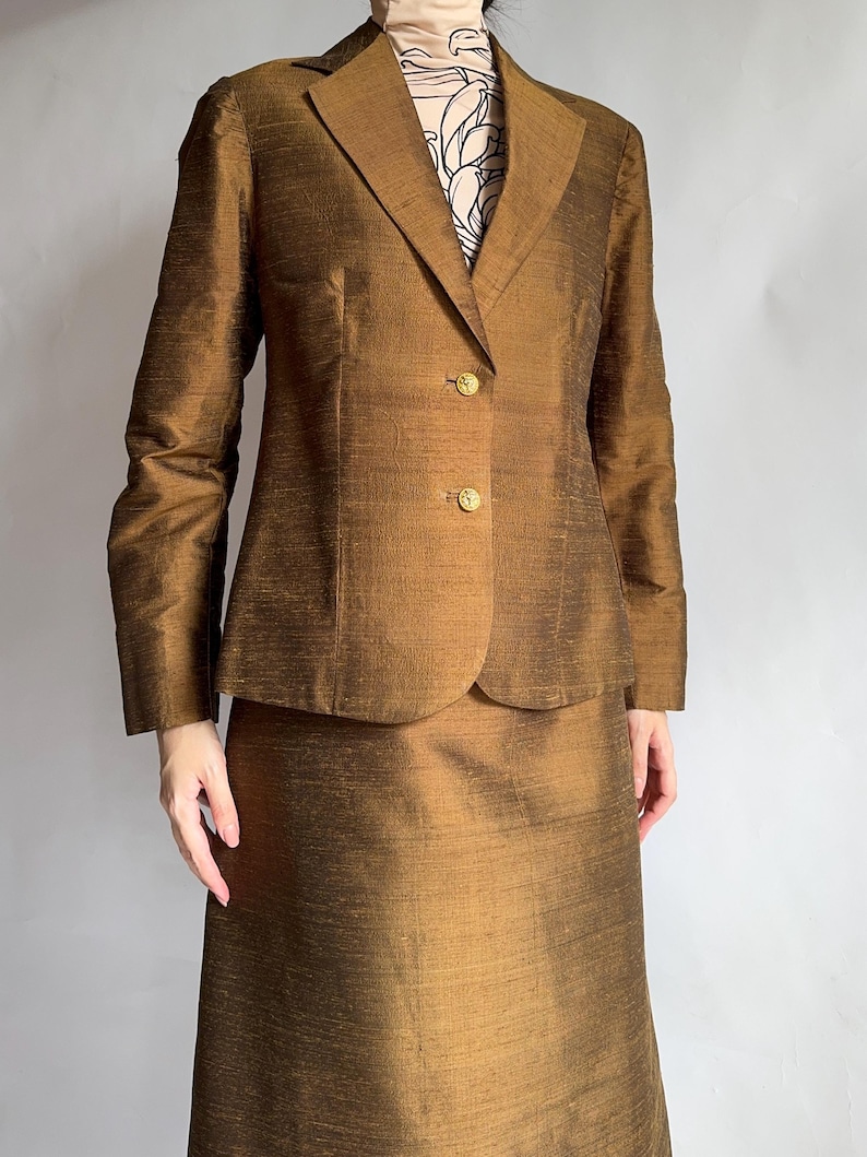 May include: A brown silk blazer and skirt suit. The blazer has a notched collar, long sleeves, and a single-breasted closure with gold buttons. The skirt is knee-length and has a straight silhouette.