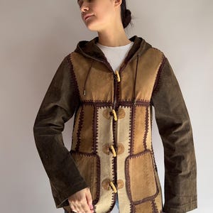 Op de afbeelding: Een bruin suède jasje met een patchwork ontwerp. De jas heeft een capuchon, knevelsluitingen en donkerbruine mouwen. De voorpanelen zijn een mix van licht en donkerbruin suède, met decoratieve stiksels.