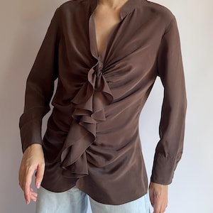 Puede incluir: Una blusa marrón de manga larga con un detalle de volantes en la parte delantera. La blusa tiene un escote en V y un diseño de nudo fruncido en la cintura. Las mangas tienen puños abotonados. La tela parece suave y sedosa.