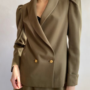 May include: A double-breasted blazer in a muted olive green colour with a wide collar and puffed sleeves. The blazer has gold buttons and a matching skirt.