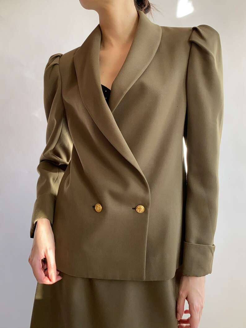 May include: A double-breasted blazer in a muted olive green colour with a wide collar and puffed sleeves. The blazer has gold buttons and a matching skirt.