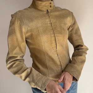 May include: A tan jacket with a snakeskin-like texture, featuring a high collar with two small metallic snap buttons, long sleeves, and a central zip closure. The garment has a structured fit and a consistent pattern across the entire surface.