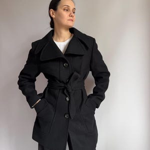 Op de afbeelding: Een zwarte wollen jas met een ceintuur in de taille en brede revers. De jas heeft twee voorzakken en knoopsluitingen. Het model draagt er een witte trui onder. Een jas in klassieke stijl.