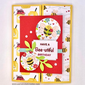 May include: A handmade birthday card with a bee theme. The card features a red center panel with the text "Have a Bee-utiful Birthday." Decorative circles with bees and flowers are included. The card is decorated with yellow and white patterned paper.