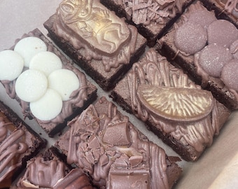 Może przedstawiać: Asortyment czekoladowych brownie z różnymi dodatkami. Niektóre są posypane białymi wiórkami czekoladowymi, wiórkami czekoladowymi i czekoladowymi cukierkami. Inne mają złote dekoracje żab i plasterków pomarańczy oraz kawałki batonów czekoladowych.