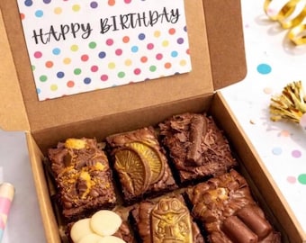 Może przedstawiać: Pudełko z dekorowanymi brownie z kartką "Happy Birthday". Brownie są posypane różnymi dekoracjami, w tym białą czekoladą, plasterkami pomarańczy i złotą żabą. Pudełko jest brązowe z kartką w kolorowe kropki.