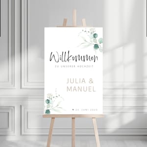 Könnte beinhalten: Ein weißes Willkommens-Schild mit grünen und weißen Blumenillustrationen. Das Schild lautet "Willkommen zu unserer Hochzeit" mit den Namen "Julia & Manuel" und dem Datum "23. Juni 2023".