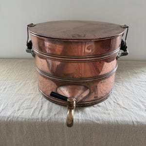 May include: A cylindrical copper vessel with a flat lid, two side handles, and a front-facing spigot featuring a black handle. The metal surface shows a warm, metallic finish with visible signs of age and use across the exterior body.