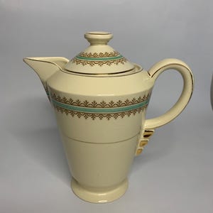 Puede incluir: Una cafetera de cerámica de color crema con un borde dorado y un diseño estampado en verde y dorado. La cafetera tiene una tapa y un asa.