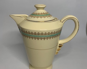 May include: A cream-colored ceramic coffee pot with a gold trim and a green and gold patterned design. The pot has a lid and a handle.