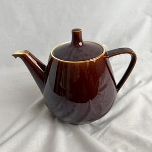 Puede incluir: Una tetera de cerámica marrón con tapa y asa. La tetera tiene un acabado brillante y un pico. La tapa tiene un pequeño pomo en la parte superior. La tetera está sobre una superficie blanca.