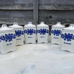 Może przedstawiać: Zestaw sześciu białych ceramicznych słoików na przyprawy z niebieskimi kwiatowymi wzorami i pasującymi pokrywkami. Każdy słoik ma inną nazwę przyprawy w niebieskich literach. Słoiki są ustawione na białej powierzchni na tle drewnianym.