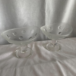 May include: Two clear glass dessert bowls with a frosted finish and circular cutouts. The bowls have a stemmed base and a shallow, oval-shaped bowl. The glass has a slightly textured appearance.