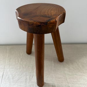 May include: A small, three-legged wooden stool with a round, thick seat featuring a natural wood grain pattern. The stool is finished in a warm, medium-brown tone, showcasing the organic texture and knots of the wood on the top surface.