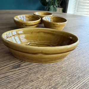 Puede incluir: Un juego de cuatro cuencos de cerámica con un esmalte marrón dorado cálido. Los cuencos varían en tamaño, con un cuenco ovalado más grande en primer plano y tres cuencos redondos más pequeños detrás. Los cuencos están colocados sobre una mesa de madera.