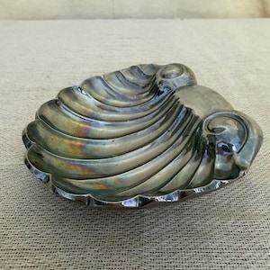 May include: A metallic, shell-shaped dish with a polished, reflective surface. The object features deep, curved ridges radiating from a smooth, rounded base, displaying iridescent, rainbow-like highlights across its dark, silver-toned finish.