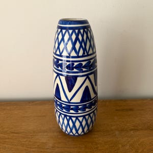 Può includere: Un vaso in ceramica blu e bianco di forma cilindrica. Il vaso presenta un motivo ripetuto di diamanti blu, chevron e disegni a forma di cuore su sfondo bianco. È esposto su una superficie di legno.