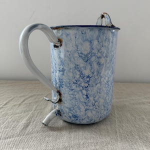 May include: A cylindrical enamel pitcher featuring a white and blue marbled pattern. The vessel includes a curved side handle, a small spout at the top rim, and a secondary small spout near the base, with visible rust at the attachment points.