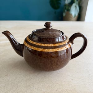 Könnte beinhalten: Eine braune Keramik-Teekanne mit rundem Körper, Ausguss und Henkel. Die Teekanne hat zwei horizontale Bänder aus beigen und braunen Streifen in der Mitte. Der Deckel hat einen kleinen Knopf oben.