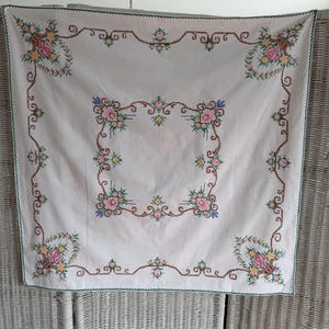 May include: A white square tablecloth with embroidered floral patterns in pink, yellow, and blue. It has a brown border and green trim. The embroidery is detailed, and the cloth appears to be made of linen or cotton.