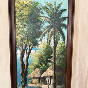 May include: A rectangular framed oil painting depicting a tropical landscape with two thatched-roof huts, a tall palm tree, and a large leafy tree near a blue body of water. A small figure stands near the huts under a light blue sky.