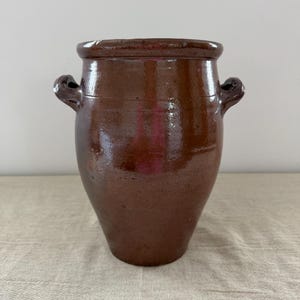 May include: A dark brown glazed ceramic vessel with a wide, rounded rim and two small, curved handles on opposite sides. The pot features a smooth, glossy finish and a tapered body that narrows towards the base, showcasing a simple, functional design.