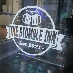 May include: A white sign with a black outline and the text "THE STUMBLE INN - Est 2021 -" with a graphic of two beer mugs. The sign is on a glass door with a refrigerator behind it. The refrigerator is filled with cans of beer.