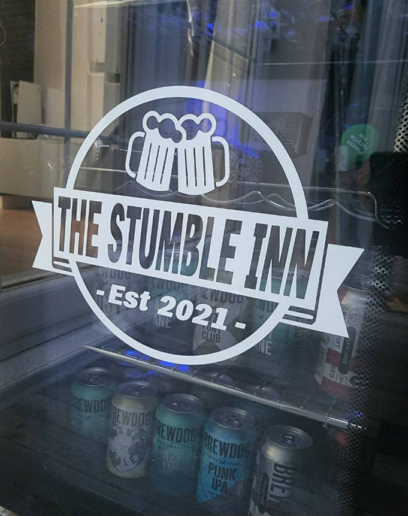 May include: A white sign with a black outline and the text "THE STUMBLE INN - Est 2021 -" with a graphic of two beer mugs. The sign is on a glass door with a refrigerator behind it. The refrigerator is filled with cans of beer.