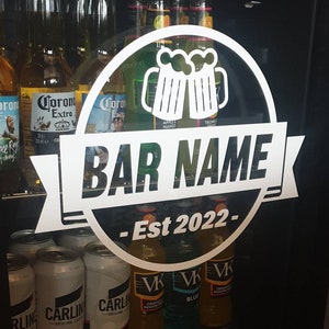 May include: A white vinyl sticker with a black circle and a white banner. The circle has two white beer mugs clinking together. The banner says "BAR NAME - Est 2022 -" in black text. The sticker is on a black refrigerator door with various beer and beverage cans and bottles.