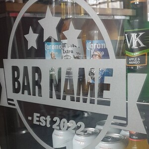 May include: A frosted glass door with a white sticker that says "BAR NAME - Est 2022" with two stars and a Corona Extra beer bottle behind it. The sticker is on a refrigerator with various beer bottles and cans, including VK Apple & Mango, Carling Original Lager, and Corona Extra.