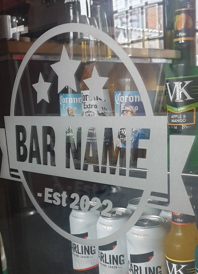 May include: A frosted glass door with a white sticker that says "BAR NAME - Est 2022" with two stars and a Corona Extra beer bottle behind it. The sticker is on a refrigerator with various beer bottles and cans, including VK Apple & Mango, Carling Original Lager, and Corona Extra.