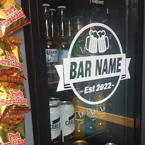 May include: A white sticker on a glass door of a refrigerator with the text "BAR NAME - Est 2022" inside a circle with a graphic of two beer mugs. The refrigerator is stocked with various beer bottles and cans.