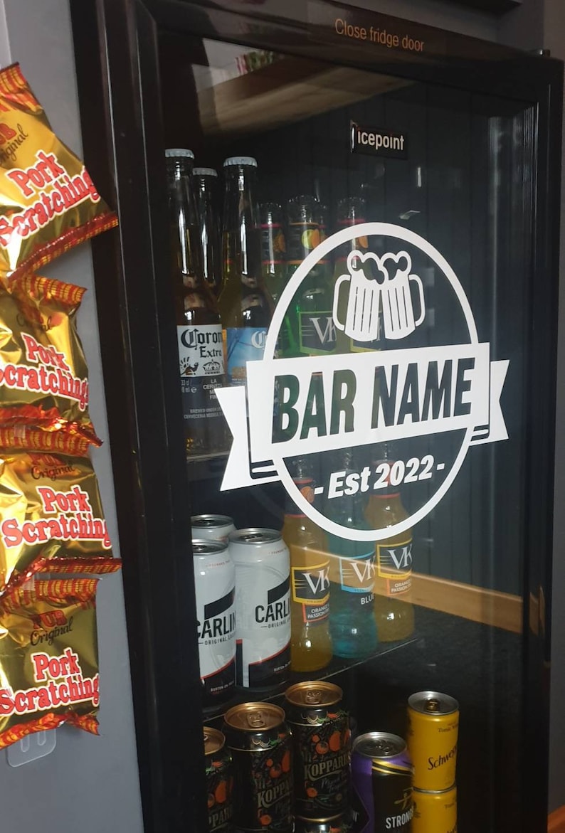 May include: A white sticker on a glass door of a refrigerator with the text "BAR NAME - Est 2022" inside a circle with a graphic of two beer mugs. The refrigerator is stocked with various beer bottles and cans.