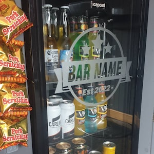 May include: A glass door refrigerator with a variety of beer and cider bottles and cans. The refrigerator has a sticker on the door that says "BAR NAME Est 2022- ".