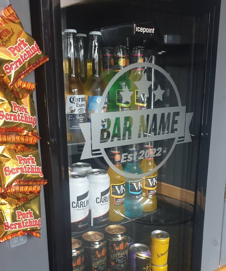 May include: A glass door refrigerator with a variety of beer and cider bottles and cans. The refrigerator has a sticker on the door that says "BAR NAME Est 2022- ".